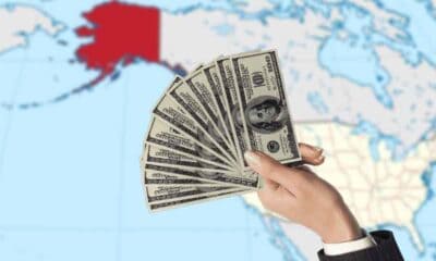 a hand offering money to Alaska on a map outlined in red