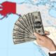 a hand offering money to Alaska on a map outlined in red