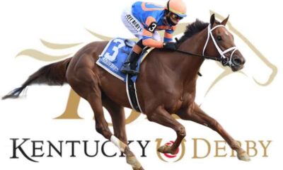 Thoroughbred horse and jockey racing at the Kentucky Derby, rider in blue and orange silks with a white bridle against Derby branding in the background.
