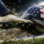 Soccer cleat kicking a muddy ball painted with the US flag on a stadium field.