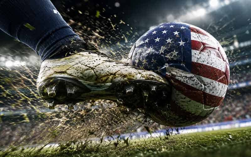 Soccer cleat kicking a muddy ball painted with the US flag on a stadium field.