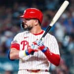 Baseball player in Philadelphia Phillies uniform prepares to swing, gripping a bat at the plate in a crowded stadium.