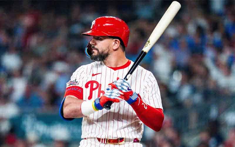 Baseball player in Philadelphia Phillies uniform prepares to swing, gripping a bat at the plate in a crowded stadium.