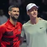 Two male tennis players pose together on a court, one in a red shirt and the other in a light gray shirt with a white cap, smiling with an arm around the other shoulder.