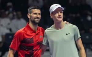 Two male tennis players pose together on a court, one in a red shirt and the other in a light gray shirt with a white cap, smiling with an arm around the other shoulder.
